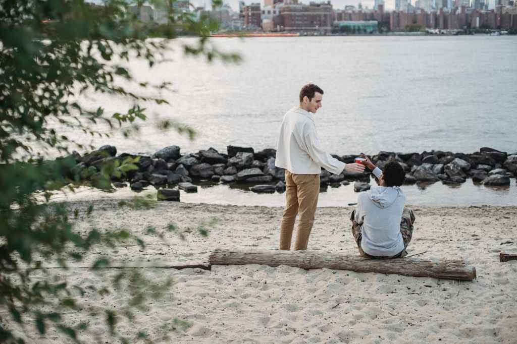young man giving paper cup to male friend sitting on seashore